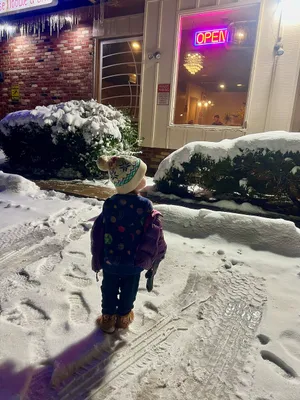 Toddler standing in a snowy parking lot at night looking into the window of an open restaurant