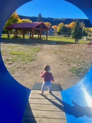 A toddler stands looking out of a tall, circular blue playground tunnel