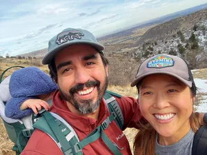 Andrew, Theresa, and a toddler passed out face-first in a hiking backpack in front of the view above the Denver foothills