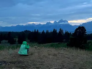 Woman in jacket squatting and taking a picture of the Tetons at sunrise