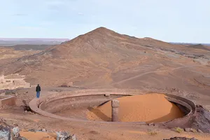 A man walking on the rim of a circular concrete pit partially filled with sand