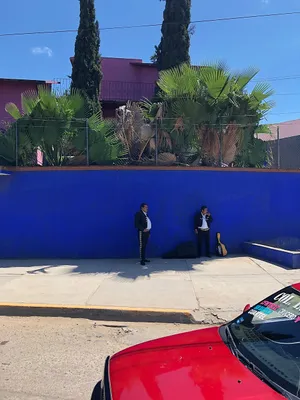Red car in foreground under mariachi members taking shade under a bright blue wall